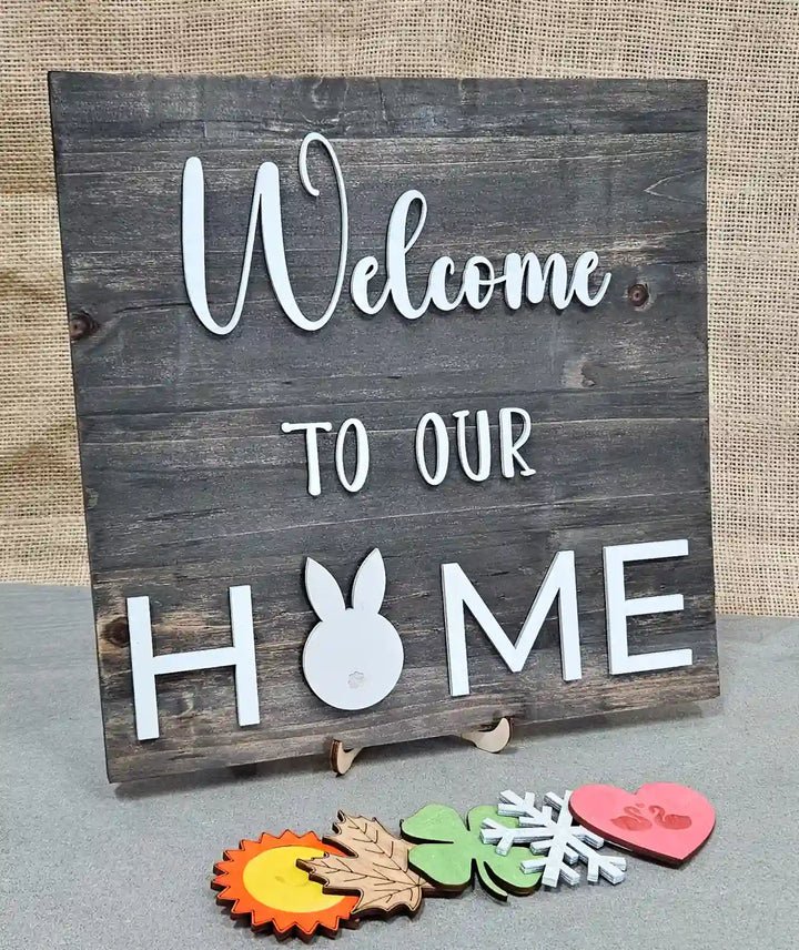 Wooden sign with a dark gray stained background and 'Welcome' in white script, 'TO OUR' in smaller white block letters, and 'H🐰ME' in larger white block letters with a white bunny silhouette replacing the 'O', displayed with five interchangeable magnetic shapes below: a sun, a maple leaf, a clover, a snowflake, and a heart with swans, against a burlap background.