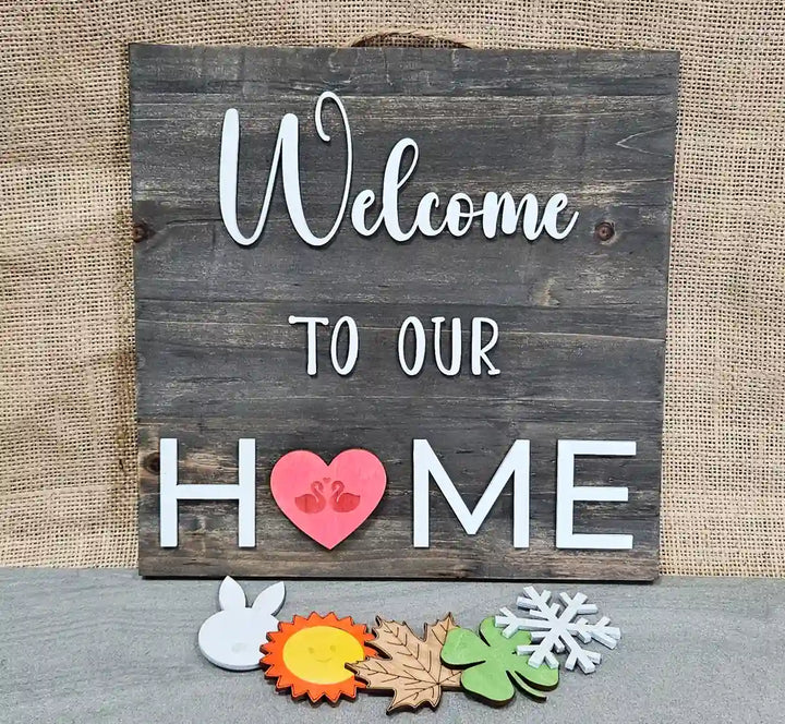 Wooden sign with a dark gray stained background and 'Welcome' in white script, 'TO OUR' in smaller white block letters, and 'H❤️ME' in larger white block letters with a pink heart containing two white swans replacing the 'O', displayed with five interchangeable magnetic shapes below: a bunny, a sun, a maple leaf, a clover, and a snowflake, against a burlap background.