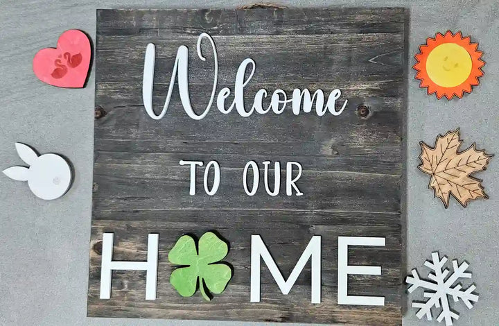 Wooden sign with a dark gray stained background and 'Welcome' in white script, 'TO OUR' in smaller white block letters, and 'H🍀ME' in larger white block letters with a green clover replacing the 'O', displayed with a pink heart with swans, a white bunny, a sun, a maple leaf, and a snowflake arranged around it, against a burlap background.