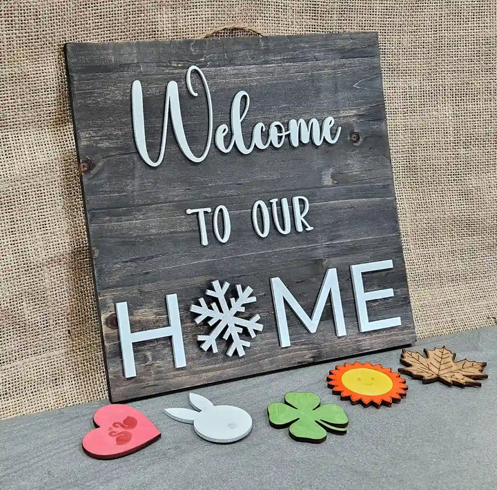 Wooden sign with a dark gray stained background and 'Welcome' in white script, 'TO OUR' in smaller white block letters, and 'H❄️ME' in larger white block letters with a white snowflake replacing the 'O', displayed with a pink heart with swans, a white bunny, a green clover, a sun, and a maple leaf arranged below it, against a burlap background.