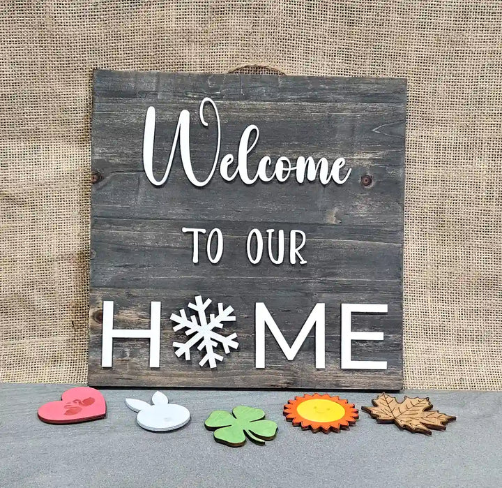 Wooden sign with a dark gray stained background and 'Welcome' in white script, 'TO OUR' in smaller white block letters, and 'H❄️ME' in larger white block letters with a white snowflake replacing the 'O', displayed with a pink heart with swans, a white bunny, a green clover, a sun, and a maple leaf arranged below it, against a burlap background.